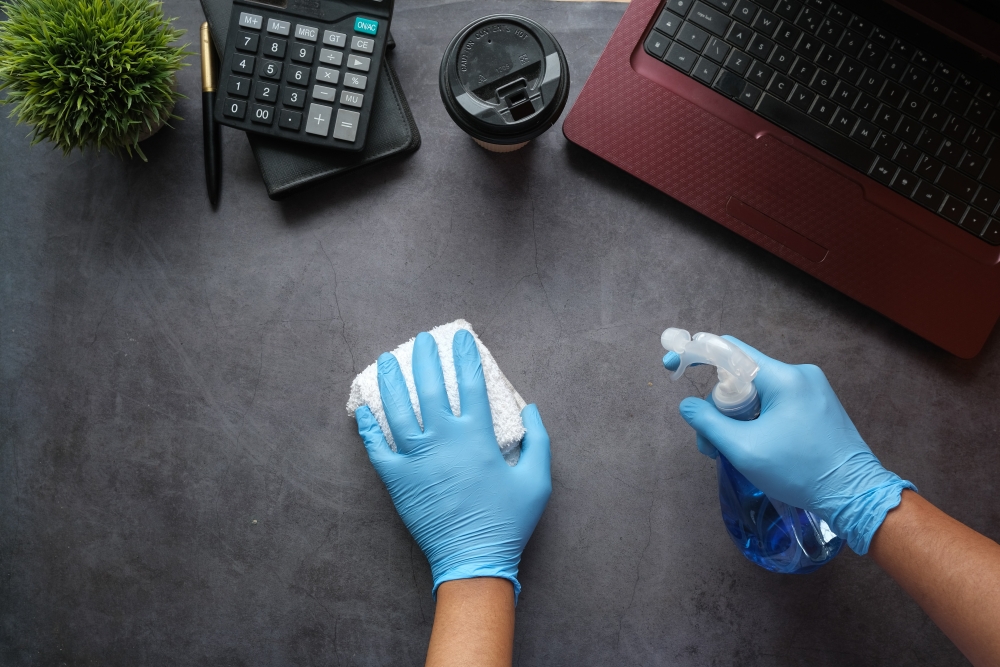 Hands wearing blue latex gloves and using cleaning supplies to clean a desk