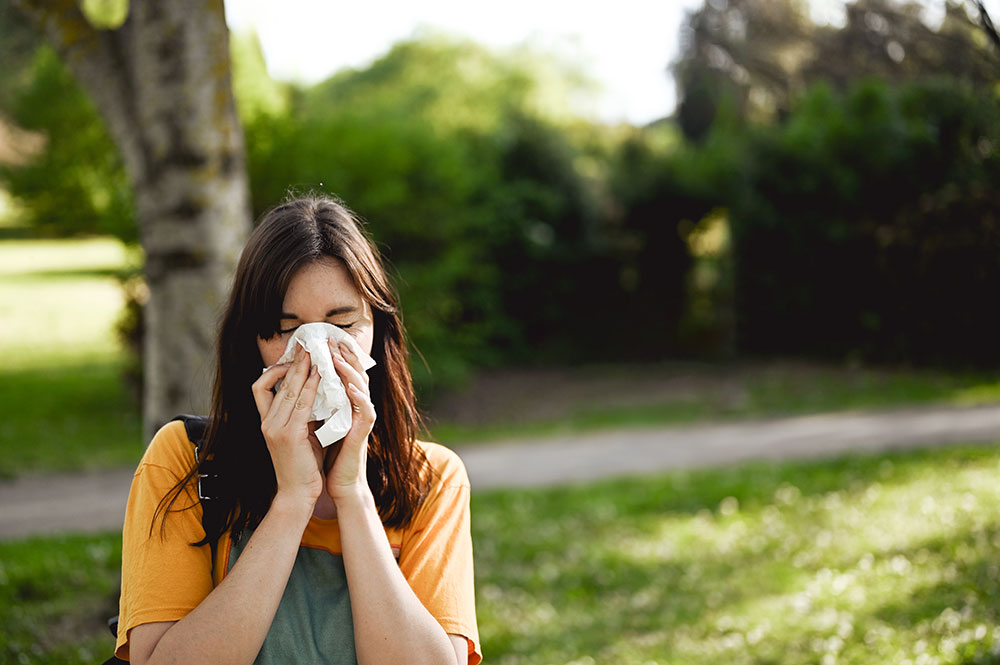 a women holding a tissue to her nose standing outside in a wooded area